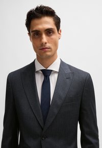 Slim-fit, dark grey suit with subtle texture, notch lapels, single button, paired with a white dress shirt and navy tie.