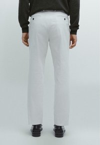 Białe spodnie proste z kieszeniami bocznymi i czarnymi guzikami, widziane z tyłu, w połączeniu z czarnymi butami i ciemnym swetrem.