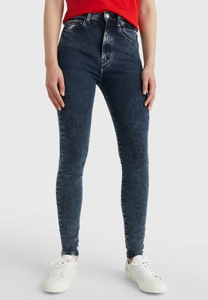 Person iført mørkeblå skinny jeans, hvide sneakers og en rød top, stående mod en ensfarvet lys baggrund.