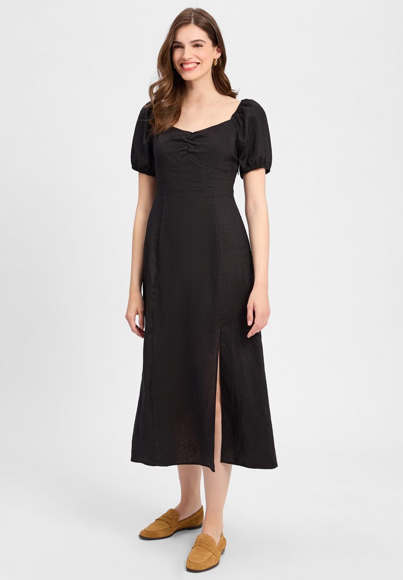 Schwarzes Leinen-Midi-Kleid mit Puffärmeln, quadratischem Ausschnitt und Mittelraffung. Mit einem Schlitz an der linken Seite. Getragen mit braunen Loafers.