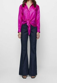 Roze satijnen overhemd met knopen en een knoop aan de voorkant, gecombineerd met donkerblauwe uitlopende jeans. De jeans hebben lichte stiksels en een gladde textuur.