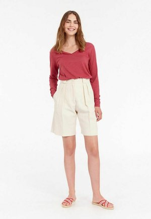 Chemise rouge à manches longues, shorts plissés couleur crème et sandales rayées rouges. Le modèle se tient devant un fond neutre, mettant en valeur l'ajustement de la tenue.