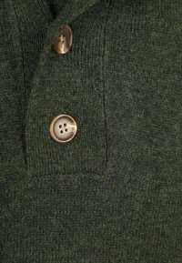 Dunkelgrüner Strickstoff mit zwei braunen, vierlochigen Knöpfen, die auf einem strukturierten Kleidungsstück genäht sind, wobei die detaillierten Nähte und Knopflöcher sichtbar sind.