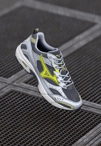 Bottines de course grises et blanches avec des accents jaunes suspendues au-dessus d'un sol en grille métallique.