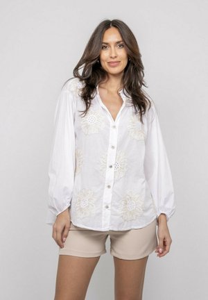 Femme aux longs cheveux bruns portant un chemisier blanc à boutons avec broderie florale et un short beige, debout devant un fond clair uni.