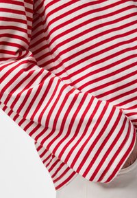 Chemise à rayures horizontales rouges et blanches à manches longues portée avec le bras plié montrant une partie de la main dans une poche blanche.