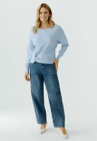 Frau steht und trägt einen hellblauen Pullover mit langen Ärmeln, blaue Jeans mit weitem Bein und weiße spitze Pumps vor einem einfarbigen hellen Hintergrund.