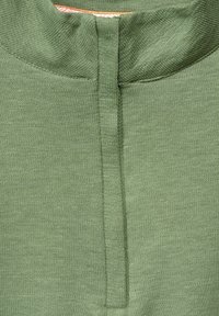 Grön bomullströja med hög krage, enkel sömmar och en kort slits framtill vid halsringningen. Mjukt, texturerat tyg.