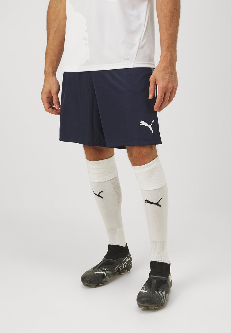 Calções de desporto azul-marinho com o logótipo branco da Puma, combinados com meias brancas com logótipo preto e sapatilhas de futebol pretas com detalhes texturizados.