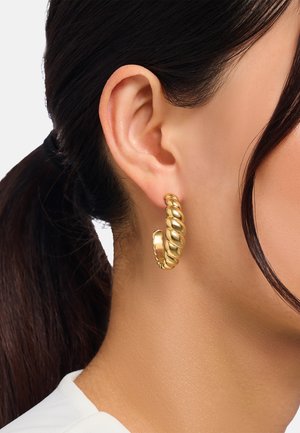 Goldene Creolen mit einem gedrehten Design, die eine glänzende Oberfläche und eine strukturierte Textur aufweisen, getragen am Ohr eines Models.