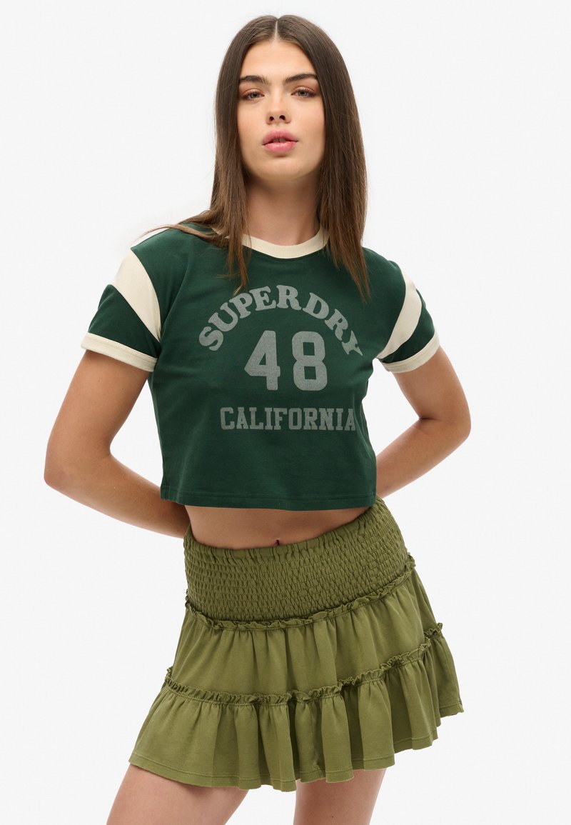 Groene crop-top met crèmekleurige mouwen, met de tekst "SUPERDRY 48 CALIFORNIA" in grijze print. Olijfgroene geraffineerde, gesmokte rok eronder.