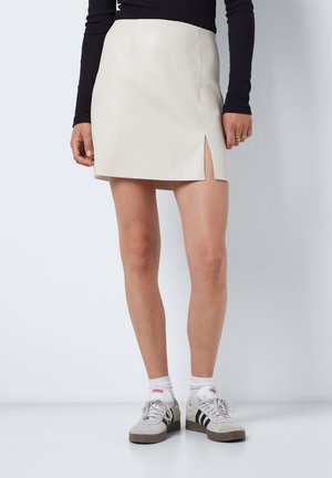 Krém színű műbőr mini szoknya oldalsó hasítékkal, sima textúrával, testhezálló szabással és minimális varrási részletekkel. Sportcipőkkel párosítva.