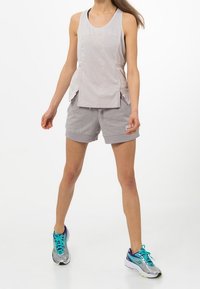 Lichtgrijs sporttanktop met zijsplits, paired met grijze shorts. Model draagt blauwe en zilveren hardloopschoenen met teal accent.