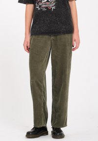 Pantaloni in corduroy verde oliva con design a gamba larga, caratterizzati da una texture a righe verticali, abbinati a stivali neri con lacci.