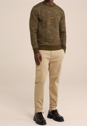 Mann trägt einen olivgrünen, strukturierten Pullover, beige Cargohosen und schwarze klobige Lederschuhe vor neutralem Hintergrund.