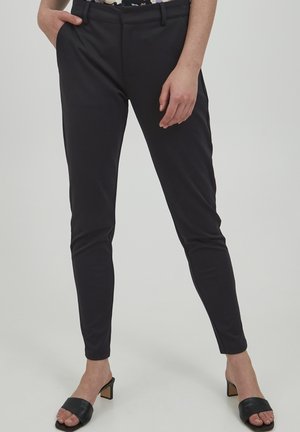 Persoon die zwarte slim-fit broek en zwarte sandalen met hakken draagt, staat met één hand in de zak en één hand ontspannen langs het lichaam.