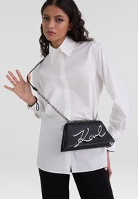 Černá kožená crossbody taška se stříbrným kovovým řetízkem, která má výrazné kurzivní logo "Karl" na přední straně a strukturovaný tvar.