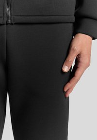 Zwarte katoenen tracksuit met een gestructureerde afwerking. Ritsdetails op de top, strakke pasvorm, hand rustend op de zijkant van de broek.