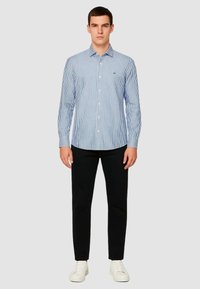 Camicia a righe in cotone blu e bianco, maniche lunghe, abbinata a pantaloni neri e sneaker bianche. Design minimalista, vestibilità sartoriale.