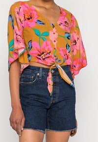 Top corto floral con flores rosas y naranjas sobre un fondo mostaza, con mangas cortas de vuelo y dobladillo atado. Combinado con shorts de mezclilla oscura.