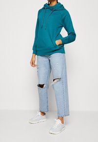 Sweat à capuche bleu sarcelle avec cordons ajustables, poche avant, associé à un jean bleu clair déchiré et des chaussures de sport blanches.