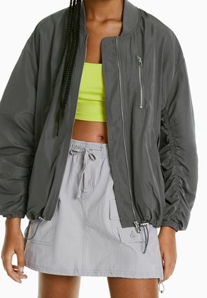 Personne portant une veste bomber grise à fermeture éclair sur un crop top vert néon et une mini-jupe gris clair à cordon avec des poches.