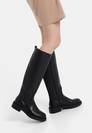 Personne portant des bottes en cuir noires montant jusqu'aux genoux avec des talons bas et un short beige, debout les jambes croisées sur un fond blanc.
