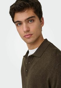 Polo marrone con colletto e tre bottoni, indossata sopra una T-shirt bianca. La texture appare morbida e leggermente spazzolata.