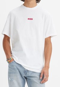 T-shirt en coton blanc avec un logo Levi's rouge central. Le t-shirt a une coupe décontractée et des manches courtes, associé à un jean bleu délavé.