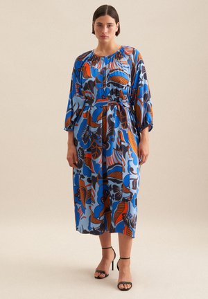 Un abito midi blu con motivo floreale, maniche ampie e scollatura arricciata, stretto in vita con una cintura di stoffa. Sandali neri con tacco.