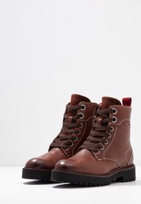 Bruna läderankelstövlar med runda tår, tjocka svarta gummisulor och bruna snören. Har en röd dragflik och kontrasterande sömmar.