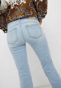 Světle modré džíny z denimu s obepínajícím střihem, které mají dvě zadní kapsy, jemné obnošení a světlou nášivku nad pravou kapsou.