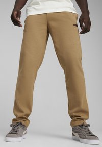 Pantaloni della tuta marroni con una vestibilità affusolata, realizzati in materiale morbido. Presentano tasche laterali e un logo discreto sulla coscia. Abbinati a sneaker grigie.