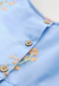 Tissu bleu clair avec des boutons marron et une broderie florale orange près d'un col orné d'un bord en dentelle rose.