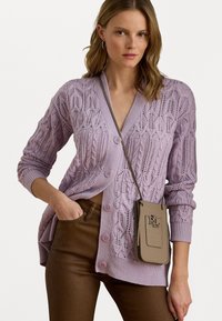 Fialový pletený cardigan s káblovými vzormi, štyri svetlofialové gombíky a rebrované manžety. Nosený cez hnedé nohavice s malou béžovou crossbody taškou.