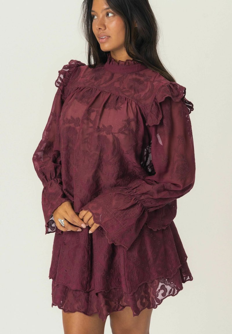 Robe en dentelle bordeaux avec col et manches volants, design superposé, motif floral et texture transparente. Présente un ourlet à étages.
