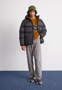 Giacca a vento nera con cappuccio, caratterizzata da un design trapuntato e un rivestimento in camouflage verde. Indossata sopra un maglione marrone e pantaloni a righe grigie.
