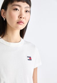 Witte katoenen t-shirt met korte mouwen, ronde halslijn en een klein, geborduurd "TOMMY JEANS" logo in blauw, rood en wit op de borst.