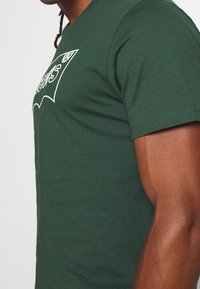 Muž ve fitted tmavě zeleném tričku s bílým textem částečně viditelným na hrudi, ukazující svalnatou ruku a boční profil.