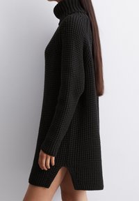 Femme dans une robe pull turtleneck en tricot noir épais avec des manches longues, se tenant de côté contre un mur blanc uni.