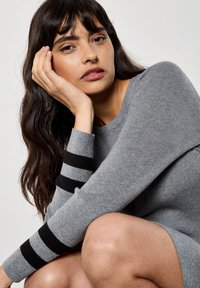 Grauer Pullover mit schwarzen gestreiften Bündchen, geripptem Textil, Rundhalsausschnitt und taillierter Passform. Das Modell sitzt mit einer Hand im Gesicht und präsentiert das Kleidungsstück.