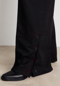 Pantalon noir avec un motif texturé, des coutures rouges le long du côté, et quatre boutons noirs au bas. Des chaussures noires sont visibles en dessous.