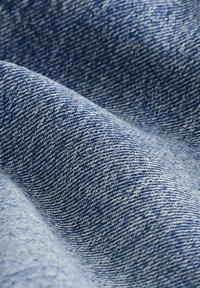 Denim tkanina u raznim nijansama plave boje, s tkanjem u riblju kost i mekim teksturom te vidljivim šavovima, koja prikazuje klasičan izgled.
