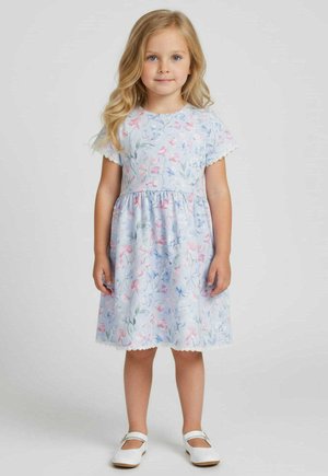 Młoda dziewczynka stojąca, ubrana w jasno-niebieską sukienkę w kwiaty z koronkowym wykończeniem i białe buty Mary Jane na jednolitym tle.