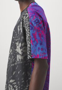 Mann trägt ein kurzärmeliges Rundhals-Shirt mit schwarzer und grauer Vorderseite sowie blau-rosa abstraktem Muster auf den Ärmeln vor weißem Hintergrund.