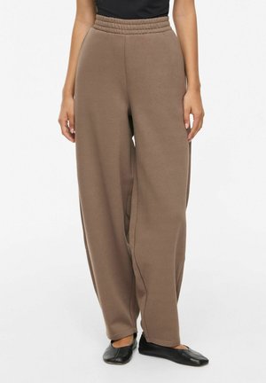 Femme portant un pantalon marron ample avec une taille élastique et des chaussures plates noires sans lacets, debout sur un fond blanc.