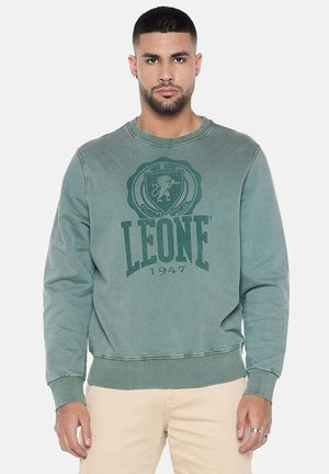 Homme portant un sweat-shirt vert à col rond avec le logo « LEONE 1947 » et un pantalon beige, debout face à l’objectif sur un fond uni.