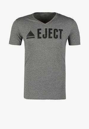 Camiseta gris de cuello en V con la palabra "EJECT" impresa, etiqueta azul. Jeans de mezclilla azul con desgaste, cadena de accesorio marrón y zapatillas grises. Mochila negra.