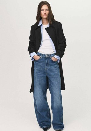 Un trench-coat noir sur un haut blanc et une chemise à boutons bleue, associé à un jean bleu à jambes larges. Design simple, coupe décontractée, pas de motifs notables.