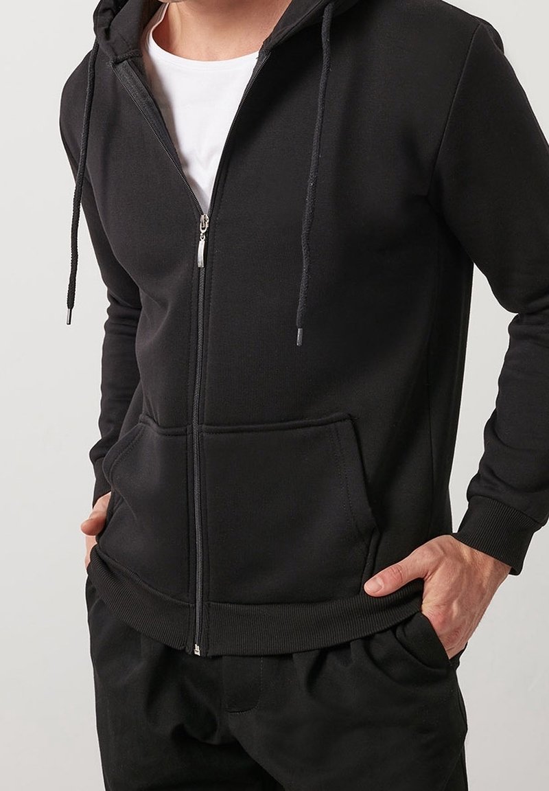 Homme portant un sweat à capuche noir zippé sur un T-shirt blanc, les mains dans les poches, sur un fond clair uni.